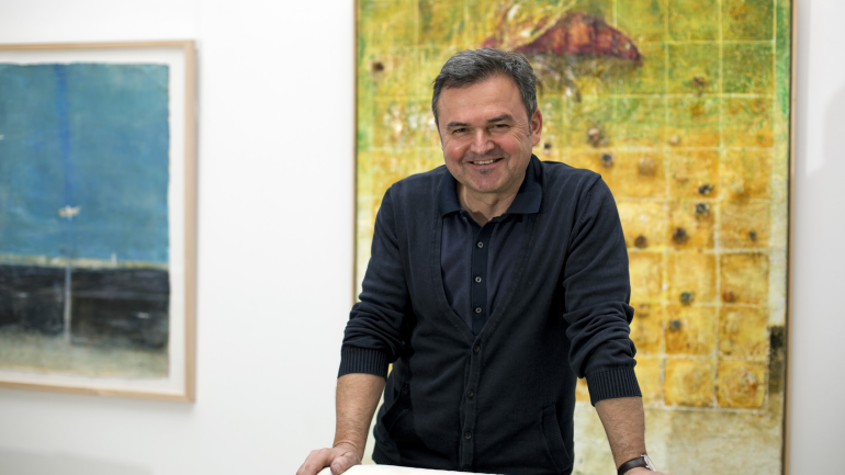 Der österreichische Künstler Manfred Leirer in seiner Galerie, vor zwei Enkaustik-Gemälden, darunter ein Werk mit Insekt und Rasterstruktur – charakteristisch für seine künstlerische Handschrift