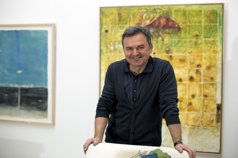 Der österreichische Künstler Manfred Leirer in seiner Galerie, vor zwei Enkaustik-Gemälden, darunter ein Werk mit Insekt und Rasterstruktur – charakteristisch für seine künstlerische Handschrift